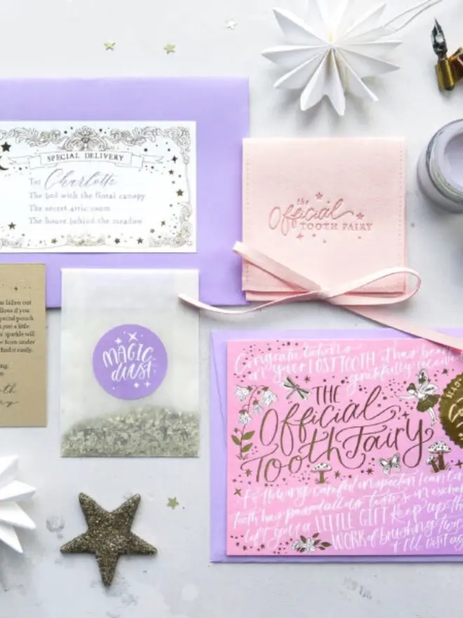 The pink set from above: addressed lilac envelope, blush pink pouch, notecard, Magic Dust and a note from the Tooth Fairy. A glitter star and white paper stars are scattered around.