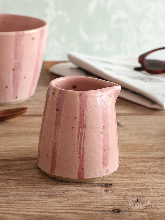 Handmade Ceramic Stoneware Small Jug in Pink Stripe sat on a wooden surface. A mug in the same style sits behind the mug, alongside a brass spoon and a newspaper with a pair of glasses on top. 