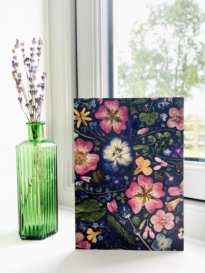 Nature-inspired greetings card with detailed pressed flower design - standing on windowsill next to green glass bottle with dried lavender - garden view