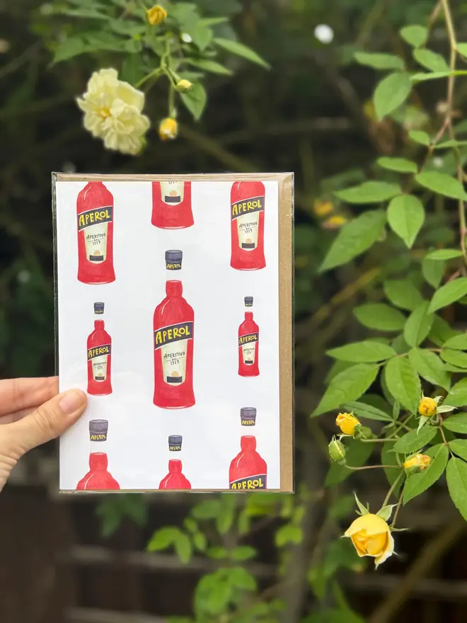 aperol spritz bottle illustrated card. card held. against leaf and flowers background.
