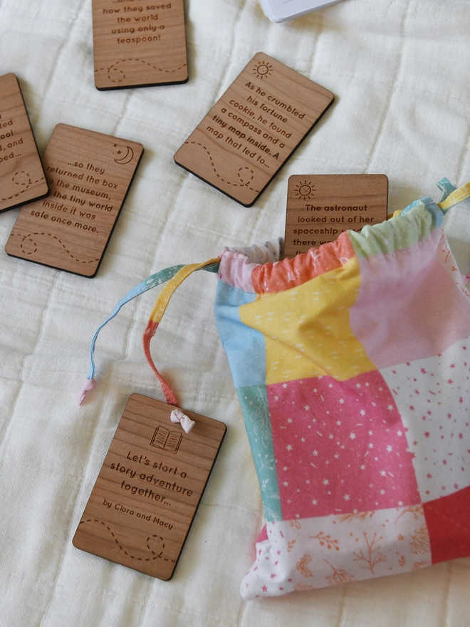 A pastel patchwork drawstring bag with wooden story cards poking out of the top. Several other wooden cards lay beside it.