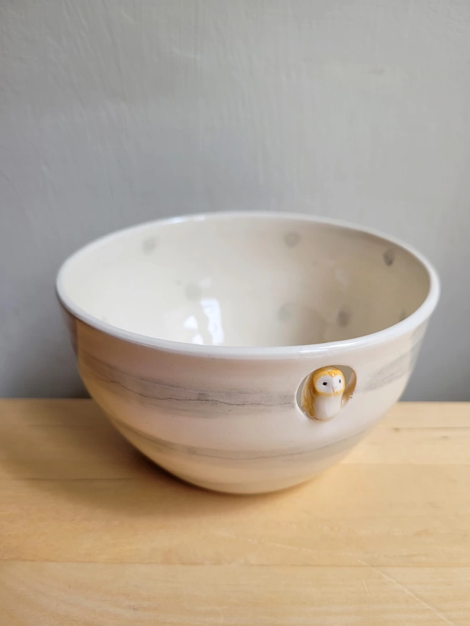 hand made ceramic owl bowl sitting on a light wooden counter the bowl is white and grey stripes with a small ceramic barn owl inside a cut out hole muted tones and detail
