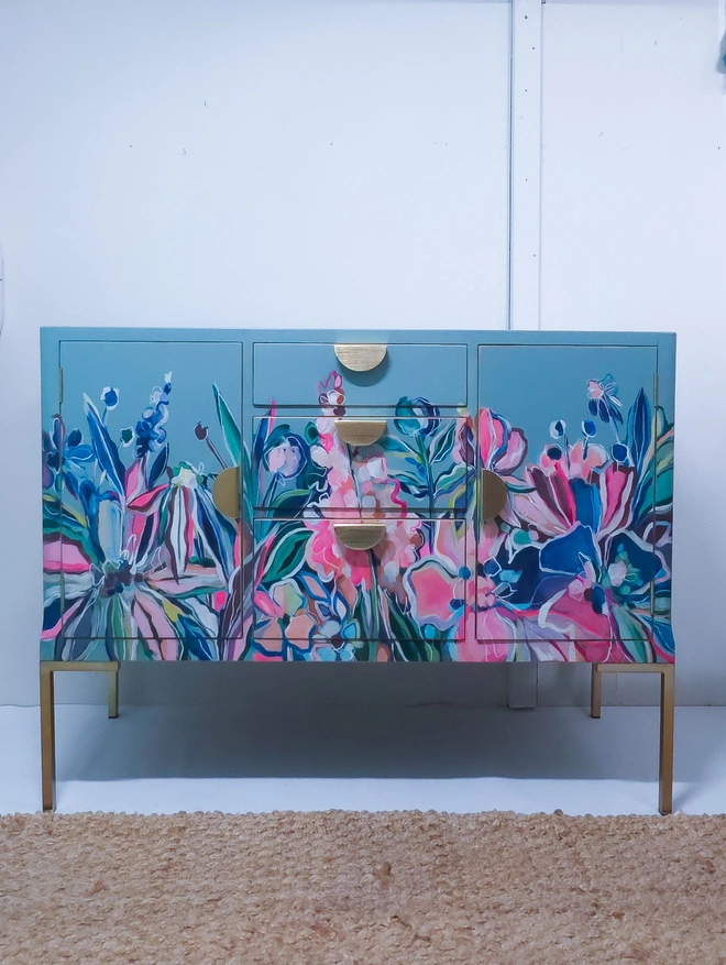 A version of ‘floral handpainted sideboard’ by chloe Kempster. Pale blue backdrop with bespoke floral design painted on top featuring peonies and daisies, the cabinet has half moon brass handles.