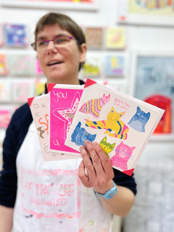Artist holding a cat card with fluro pink blue & yellow cats with curious stares & funny poses 