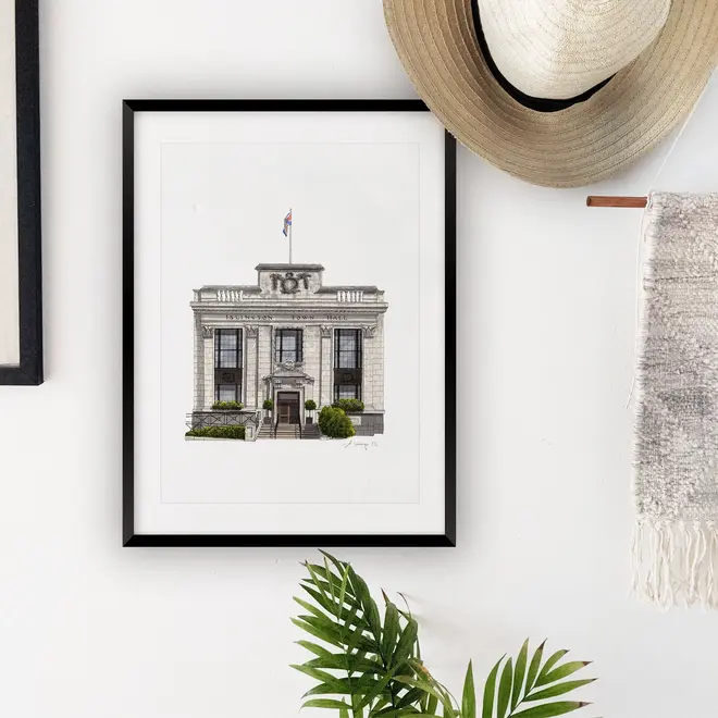 Islington Town Hall Giclée Print (Unframed), a print of a town hall sitting in a black frame against a white backdrop. It is accompanied by a hat and a plant. 