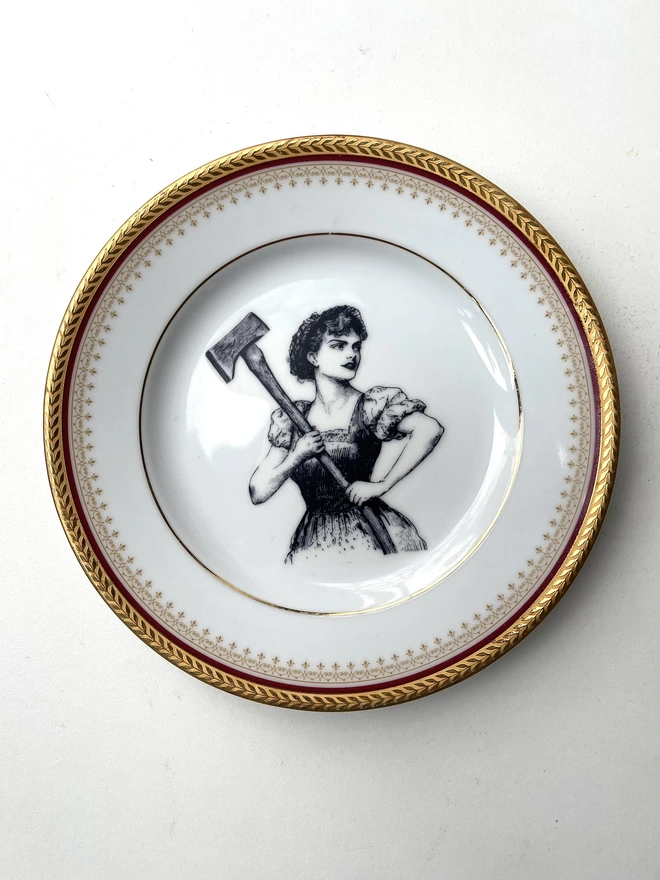 a vintage plate with a really ornate pretty border has a vintage black and white illustration of a beautiful girl in peasant clothes wielding an axe