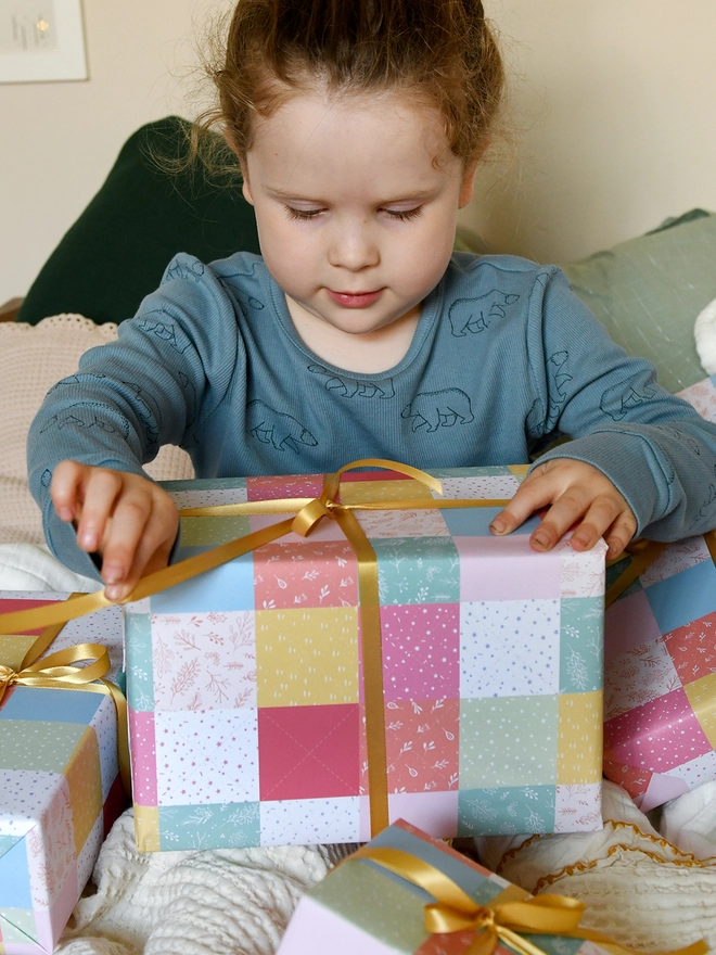 A young child wearing blue pyjamas is holding a pile of gifts wrapped in gift wrap with a design of pastel patchwork quilt design.