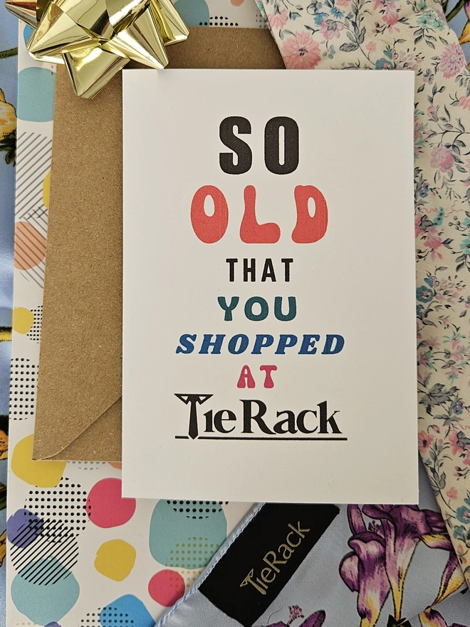 A photograph of the front of a humorous A6 greeting card illustrated using brightly coloured text and the retro shop logo. The text says so old you shopped at Tie Rack with a vintage Tie Rack scarf and floral tie 