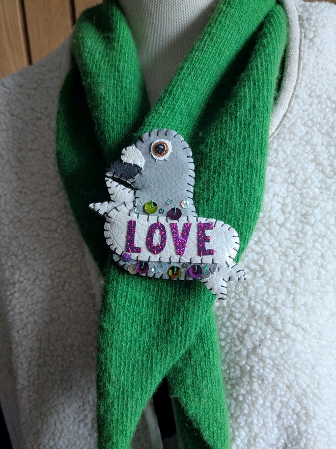 Hand-stitched white and grey faux leather pigeon brooch with purple glitter ‘LOVE’ lettering and colourful sequin details, pinned to a bright green scarf