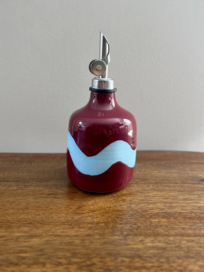 burgundy oil bottle with light blue wave pattern on a wooden table, oil bottle has straight sides and a glossy glaze contrasted with a matt wave design