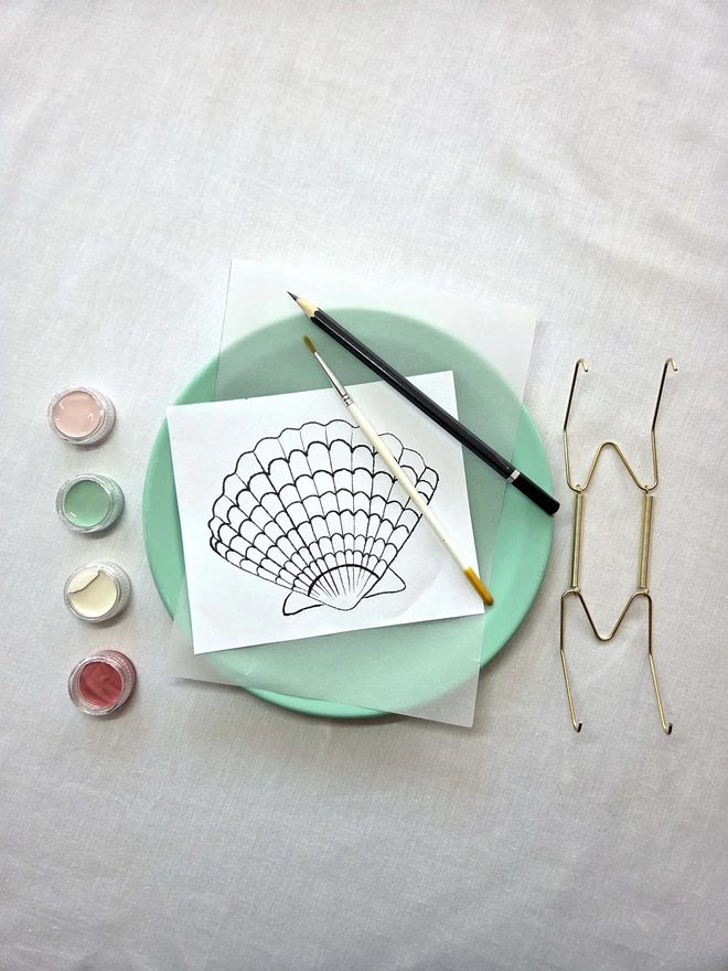 Contents of the shell plate painting kit on a white cloth - plate, design template, brush, pencil, paints and wall hook. 