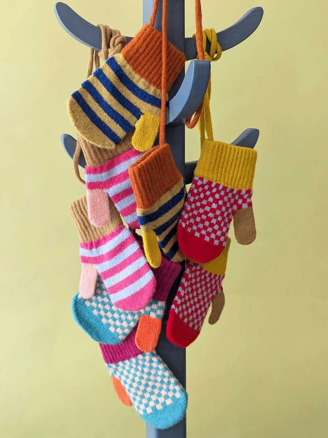 A Selection Of Kids Mittens On A String Hanging On A Coat Stand In Front Of  A Green Background