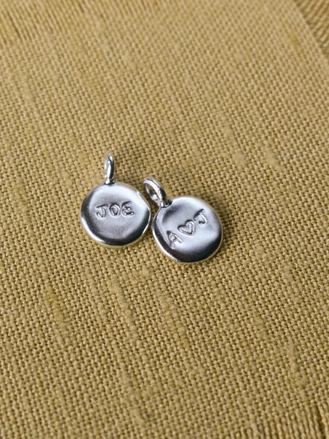 two shiny silver coin shaped pendants, one with 'JOE' and the other with 'A HEART J' in hand engraved. they lay on a woven yellow fabric background.