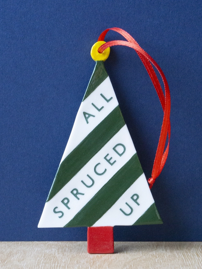 Ceramic porcelain Christmas decoration with All Spruced Up written on the side. A white tree-shaped Christmas decoration with green lettering, decorated with green stripes and with a red satin ribbon.