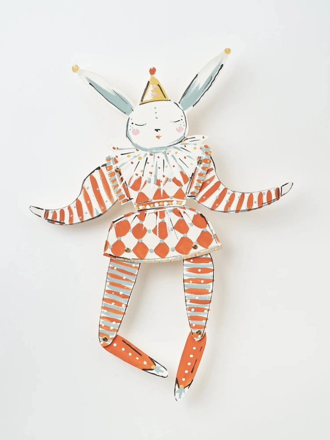 Pierrot Wall Puppet. a flat lay product shot of the pierrot wall puppet on a plain white background, showing its jointed wooden limbs painted in white, burnt orange and pale blue tones. the hare has a closed-eye, serene expression, wearing a harlequin-style outfit with a diamond-patterned skirt, striped tights and a ruffled collar, topped with a small pointed party hat. brass fasteners at the shoulders, hips and knees allow the limbs to move, giving the puppet a playful, articulated character.