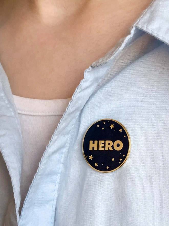 A navy blue and gold enamel pin badge with a starry design and the word "Hero" is pinned to a blue shirt.