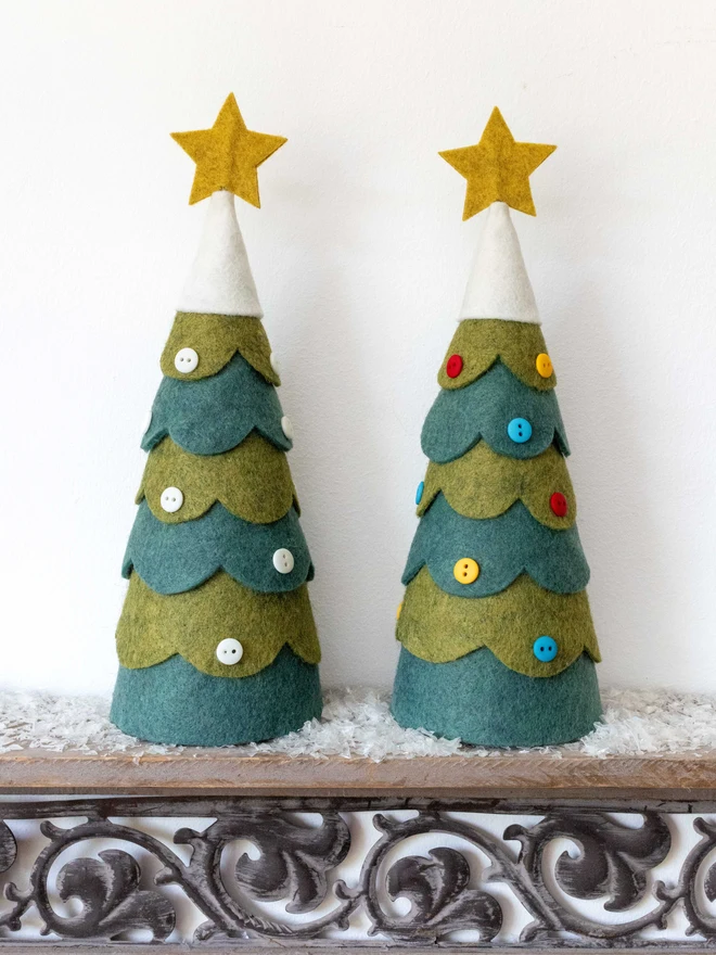 A pair of felt Christmas tree decorations. Each is decorated with buttons, one with all white, the other with red, yellow and blue. The tree has a snowy felt top adorned with a star.