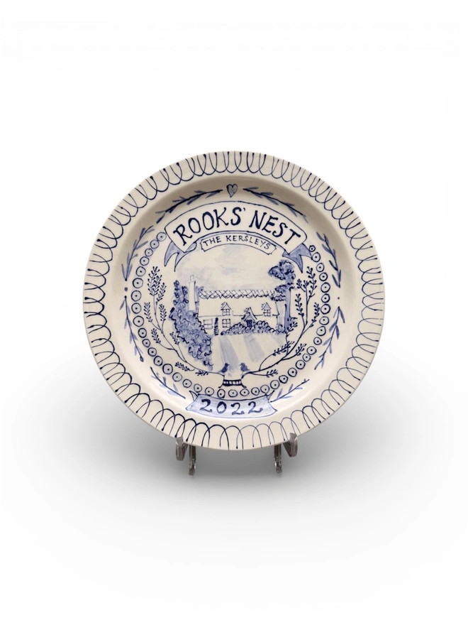 Large plate, hand painted portrait of a house cottage with botanicals around the inside rim and the name "Rooks Nest" above in hand-written capitals