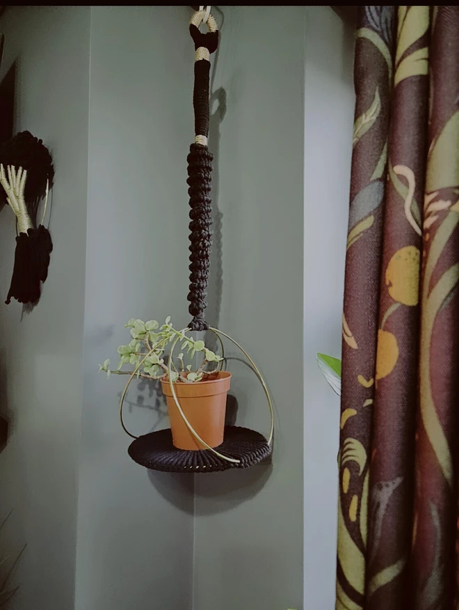 A handmade macrame plant hanger made with gold hoops and black cotton featuring gold accents and allows for a small indoor houseplant to sit on the woven base. The background is a warm home setting.