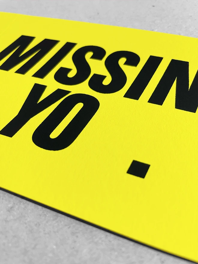 Close up of this landscape screen printed card reading Missing You. Focussed on the gap where the letter u should be.