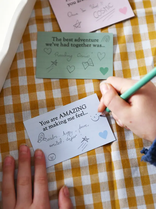 A child’s hands are shown writing on a light blue prompt card that says “You are AMAZING at making me feel…” using a green pencil. Other completed cards with handwritten messages are visible nearby on a yellow and white gingham tablecloth, including one that says “The best adventure we’ve had together was…” This scene captures a child filling out a sentimental Father’s Day gift.