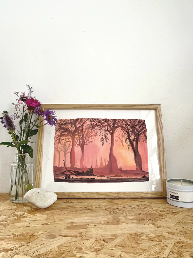 A print featuring a painted landscape illustration of a sunset through a forest with the silhouettes of trees and monkeys, in a frame next to some flowers, a rock and a candle