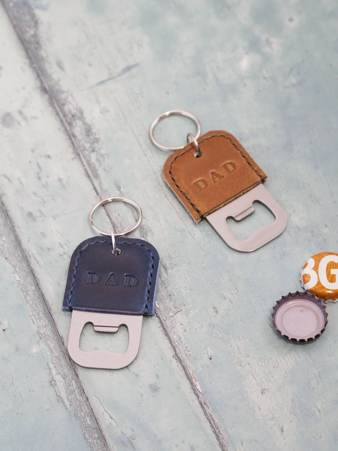 Personalised leather bottle opener key ring. Two handmade mini leather bottle openers sit with some open bottle caps. One is hand dyed dark blue, the other tan brown. Both are stamped with the name DAD.