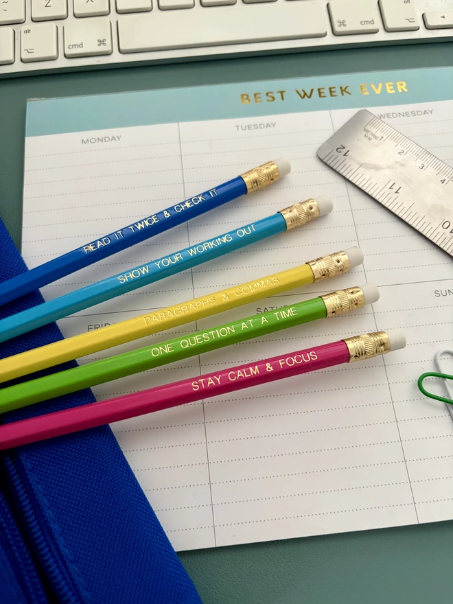 Exam essentials pencil set - Five colourful motivational exam pencils with gold foil lettering are styled on a pastel desk setup with stationery and a weekly planner.