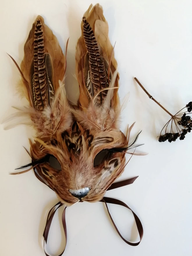 A luxury brown hare masquerade mask laying flat next to dried flowers