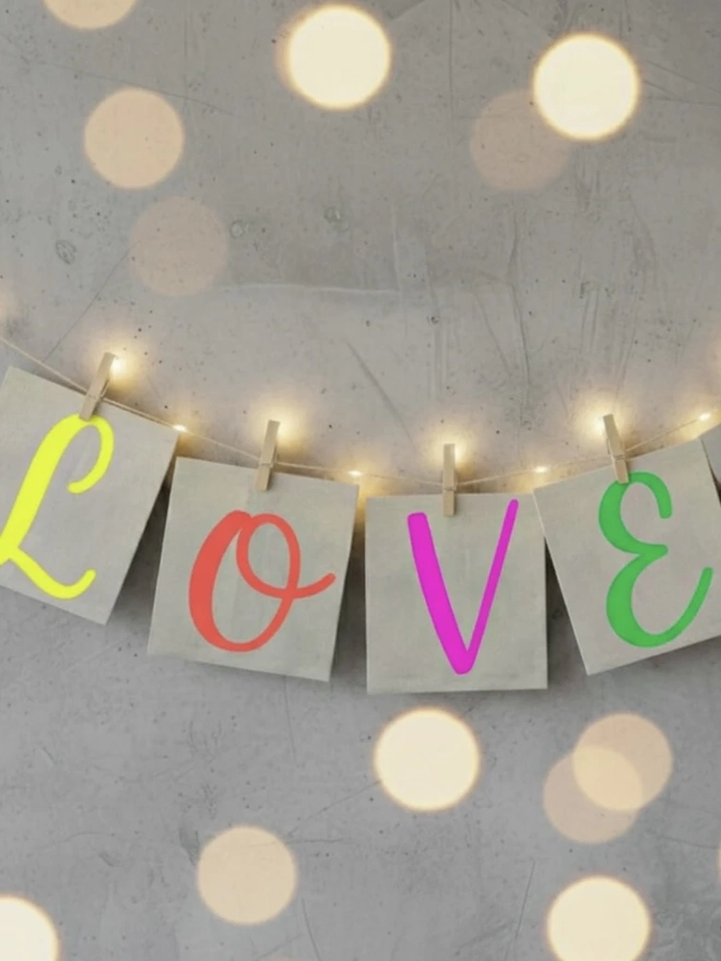 Hand Painted Alphabet Initials Linen Table Napkins, a group of colourful initial napkins hanging from string against a grey wall. It is accompanied by sparkling lights. 