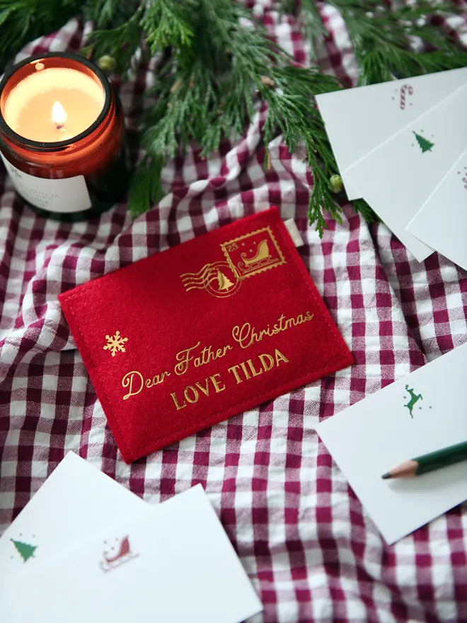 Red felt personalised Christmas envelope reading "Dear Father Christmas, Love Tilda," lying on a red gingham cloth with festive notecards, greenery, and a lit candle.