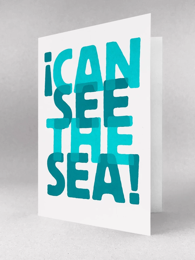 I can see the sea, screenprinted words overlapped in two turquoise blues, on a white card. It is sat in a light grey studio set, slightly open.