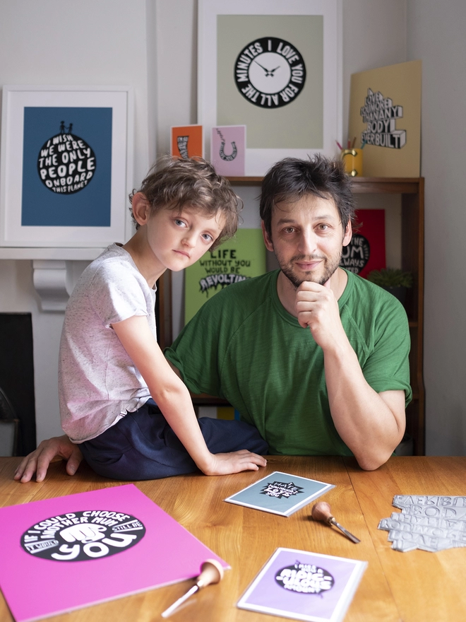 A Father and young son sit at a table, the boy is sitting on the table, they are both looking at the camera, surrounded by their Woodism art prints on the table and the wall behind them