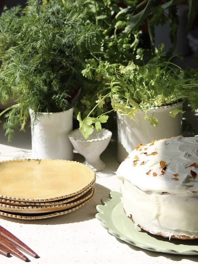 a cake shown on a sage green daisy dinner plate with a stack of scalloped edge saide plates in mustard colour. Herbs in white plant pots in background 