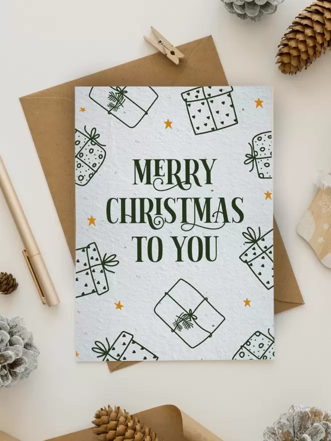 Christmas Card with 'Merry Christmas To You' with presents around the edge of the card. The card is surrounded by pine cones, a pen and a small peg