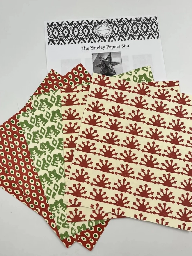 Colourful Star Kit  A Yateley Papers star kit laid out with an instruction sheet and square sheets of patterned paper in red and green prints on cream. The block-printed designs include red spot, green leaf and red sunburst patterns for folding a decorative paper star.