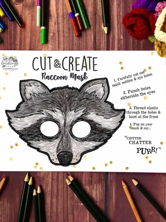 Cut And Create Forest Animals Children’s Masks Kit, an illustrated racoon face, surrounded by coloured pencils on a dark wood background.
