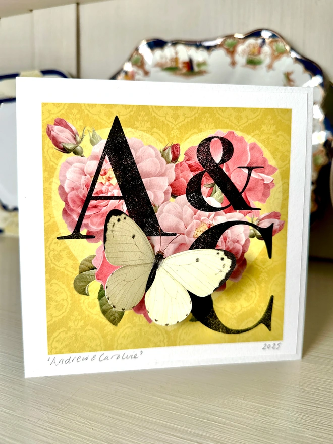 A hand made Golden wedding anniversary card featuring a heart made of deep pink roses on a golden background with letters and initials of the couple. A white paper cut three dimensional butterfly sits on the heart. 