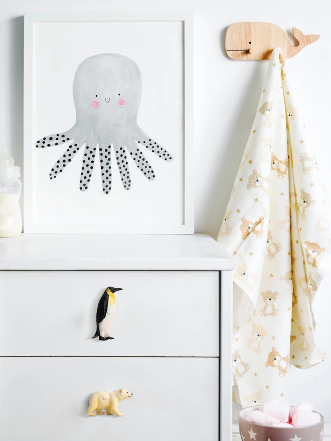 A white chest of drawers with two animal drawer knobs on it. An emperor penguin drawer knob is on the first drawer, a polar bear drawer knob is on the second drawer. Above the chest of drawers there is a watercolour cartoon like print of an octopus. To the right of the print there is a wooden hook in the shape of a whale, a cloth hangs on the hook. The animal knobs are made of plastic and made by Candy Queen Designs.