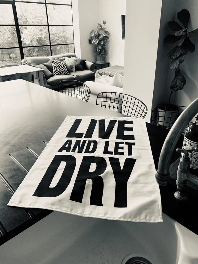 London Drying Live and Let Dry black screen printed text on white tea towel laying on sink/kitchen counter with living room in background