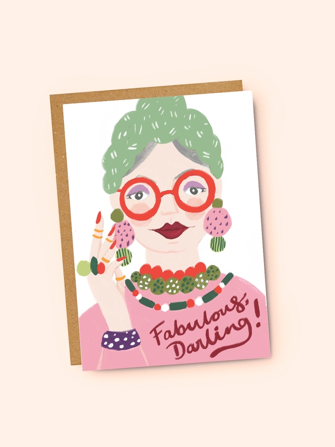 A flat lay of a greeting card that reads 'Fabulous Darling!' with an illustration of a glamorous older woman, wearing bold red glasses, and lipstick, with lots of colourful jewellery.