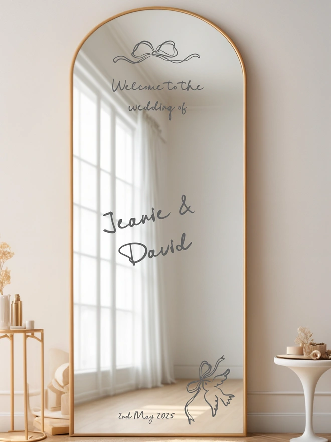 Large Mirror leaning against a white wall with a table next to it, on the mirror in black lettering is the wording Welcome to the wedding of Jeanie and David and the wedding date, at the top of the mirror is a whimiscal bow and on the botton right of the mirror is a dove carrying a bow