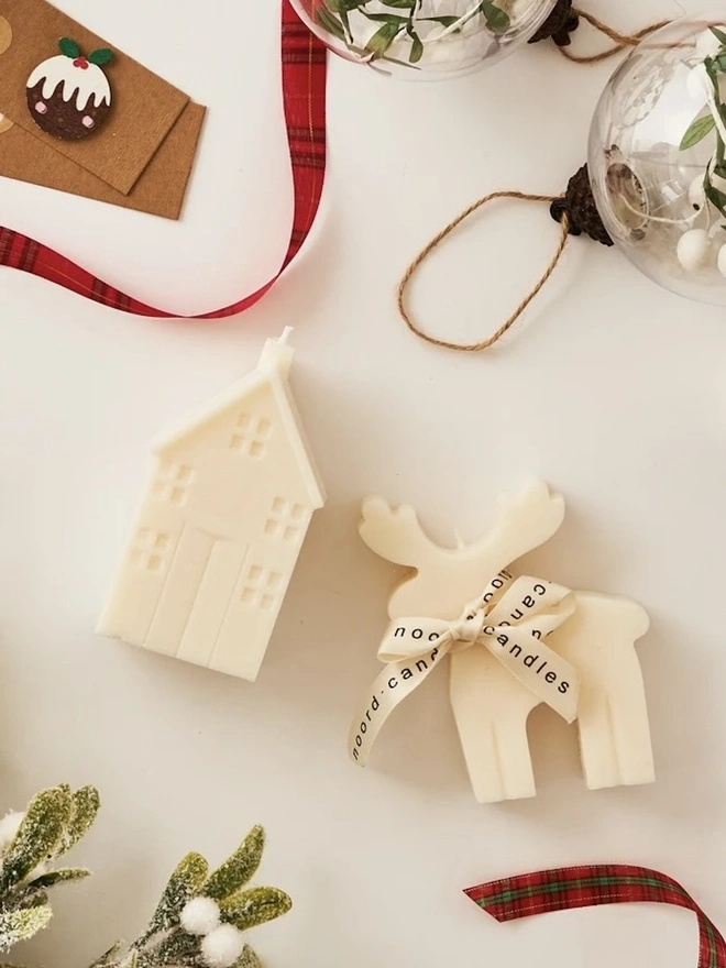 Reindeer Sculpture Candle. A flat lay of a cream reindeer candle with a branded ribbon and a matching house candle, surrounded by tartan ribbon, gift tags, frosted foliage and glass baubles. The Scandi-style Christmas candles make a charming stocking filler or Secret Santa gift.