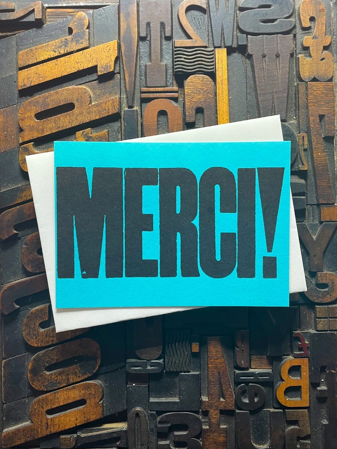 A bright turquoise thick duplex card with the word "MERCI!" printed in bold, black letters lies on a surface covered with various letterpress blocks.