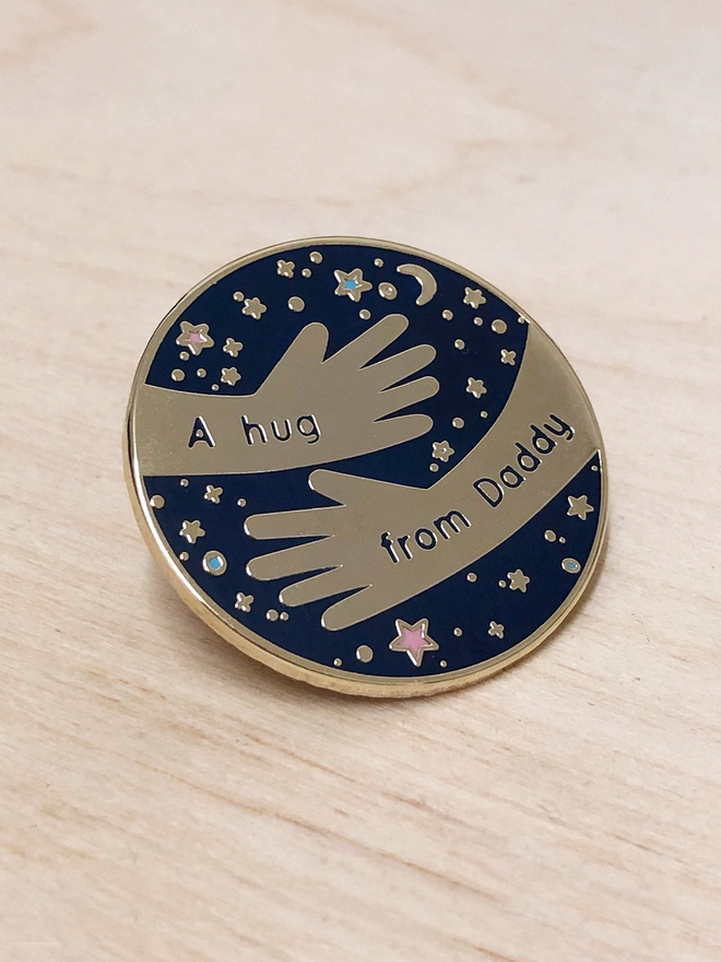 A navy blue and gold enamel pin badge with a hugging arms design and the words "A hug from Daddy" is resting on a wooden table.