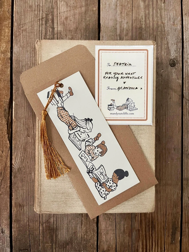 a photograph of a traditional card bookmark illustrated with a vintage children's book style drawing of kids reading books. The bookmark is cream, ochre and sepia in colour, the tassel is a golden brown. Laid on a kraft envelope and there is also a gift giving label, or bookplate sticker making it a lovely alternative to a greeting card