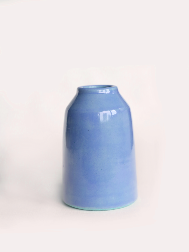 A small handmade ceramic bud vase in glossy pacific blue glaze, with a rounded bottle shape and narrow neck, photographed against a plain white background