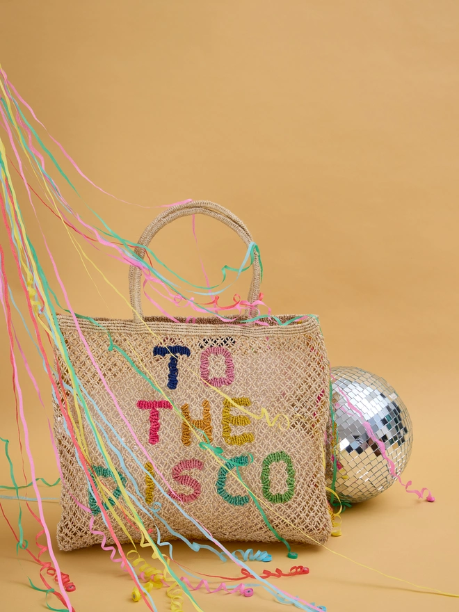 A woven bag with "To the Disco" in colorful letters is surrounded by colorful streamers on a beige background. A disco ball sits to the right, adding a festive vibe.