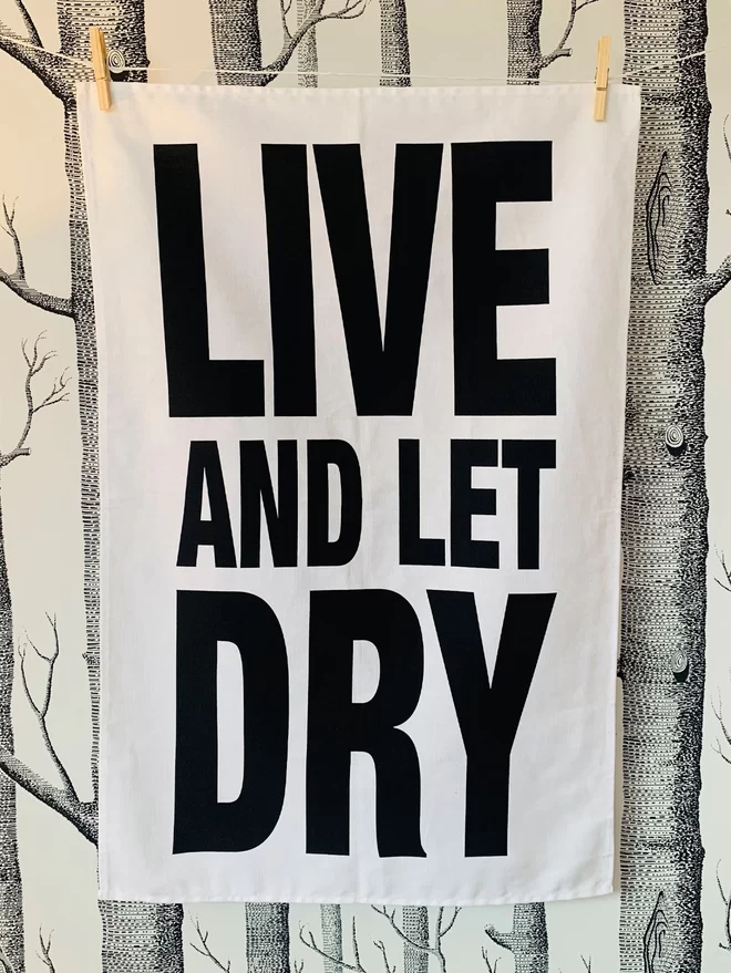 London Drying Live and Let Dry in black text on white tea towel hung washing line style in front of a black and white trees/woods pattern wallpaper