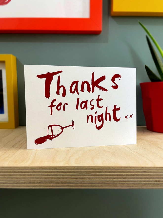 Thanks for Last night in a handwritten scrawl is screenprinted across this greetings card in claret red, with a knocked over wine glass besides. The card is stood on a plywwod shelf with hints of other framed pictures around and a plant to one side. 